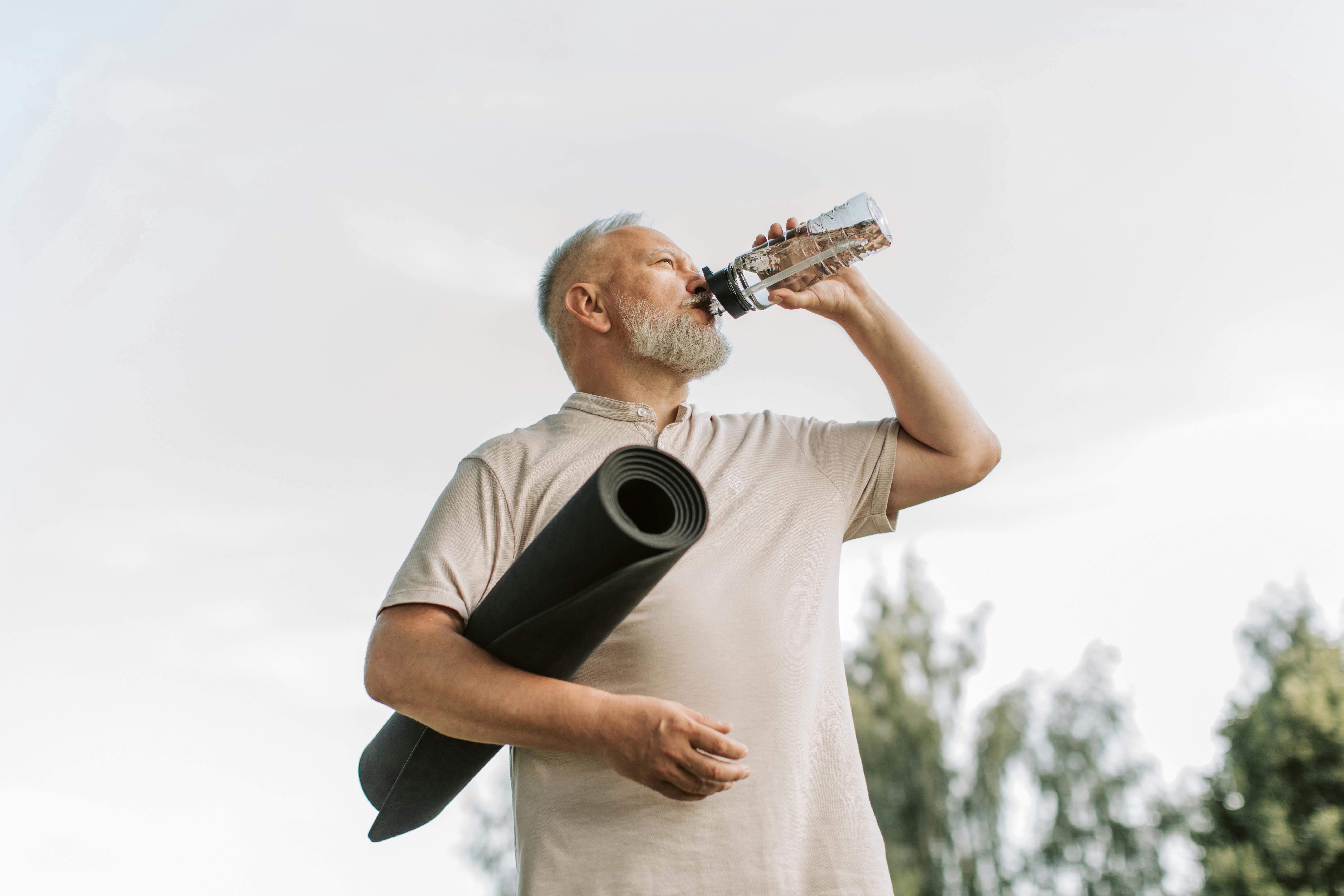 Ein junger Mann trinkt aus einer Trinkflasche. Unter seinem Arm klemmt eine Gymnastikmatte.