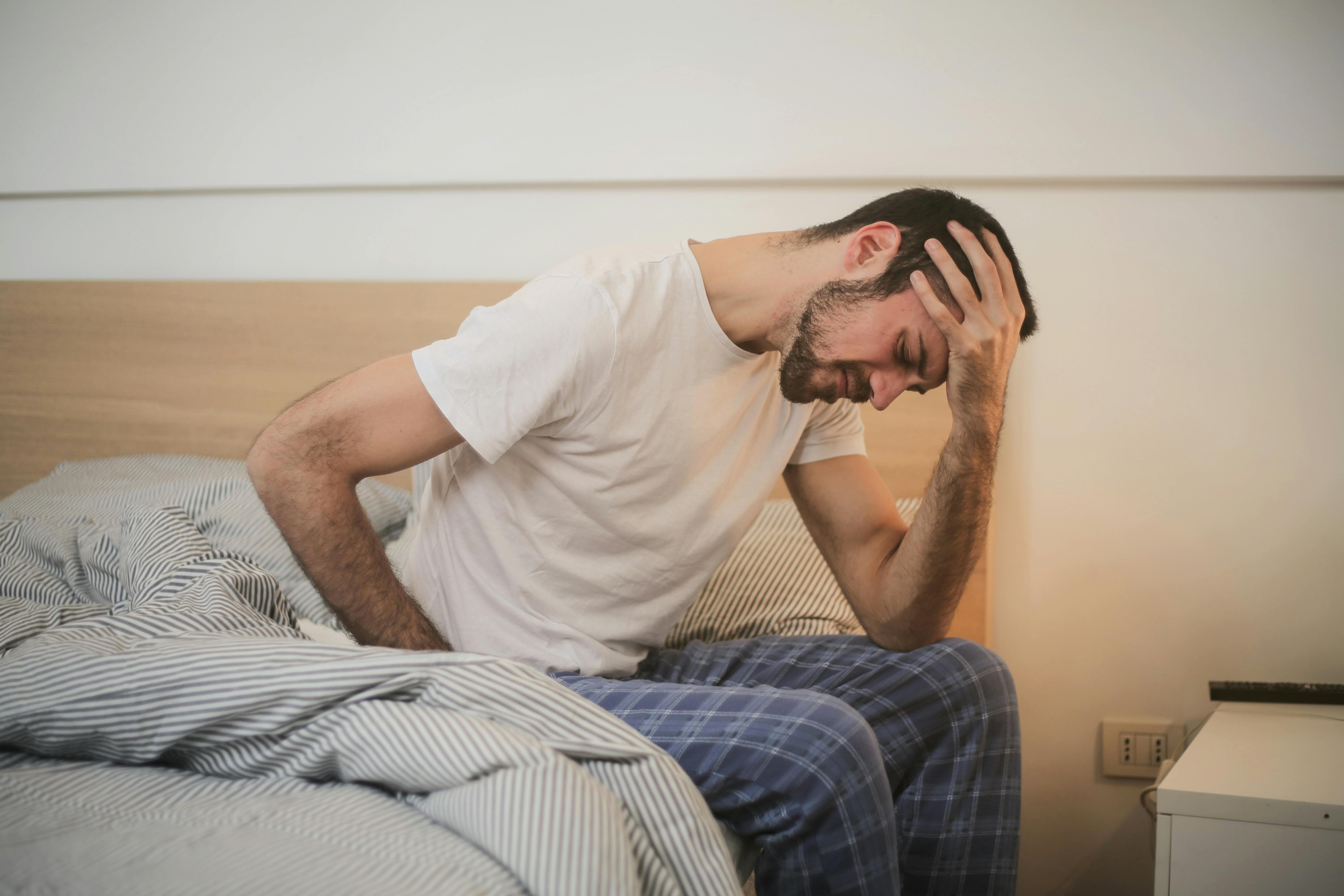 Eine Person, die müde und ausgelaugt wirkt, sitzt auf der Bettkante und stützt den Kopf in die Hände.