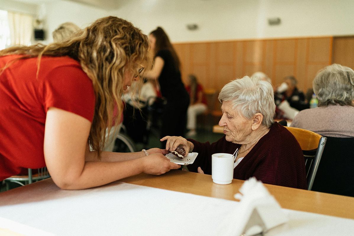 Eine Pflegefachkraft reicht im Speisesaal einer Pflegeeinrichtung einer älteren Frau ein Stück Kuchen an.