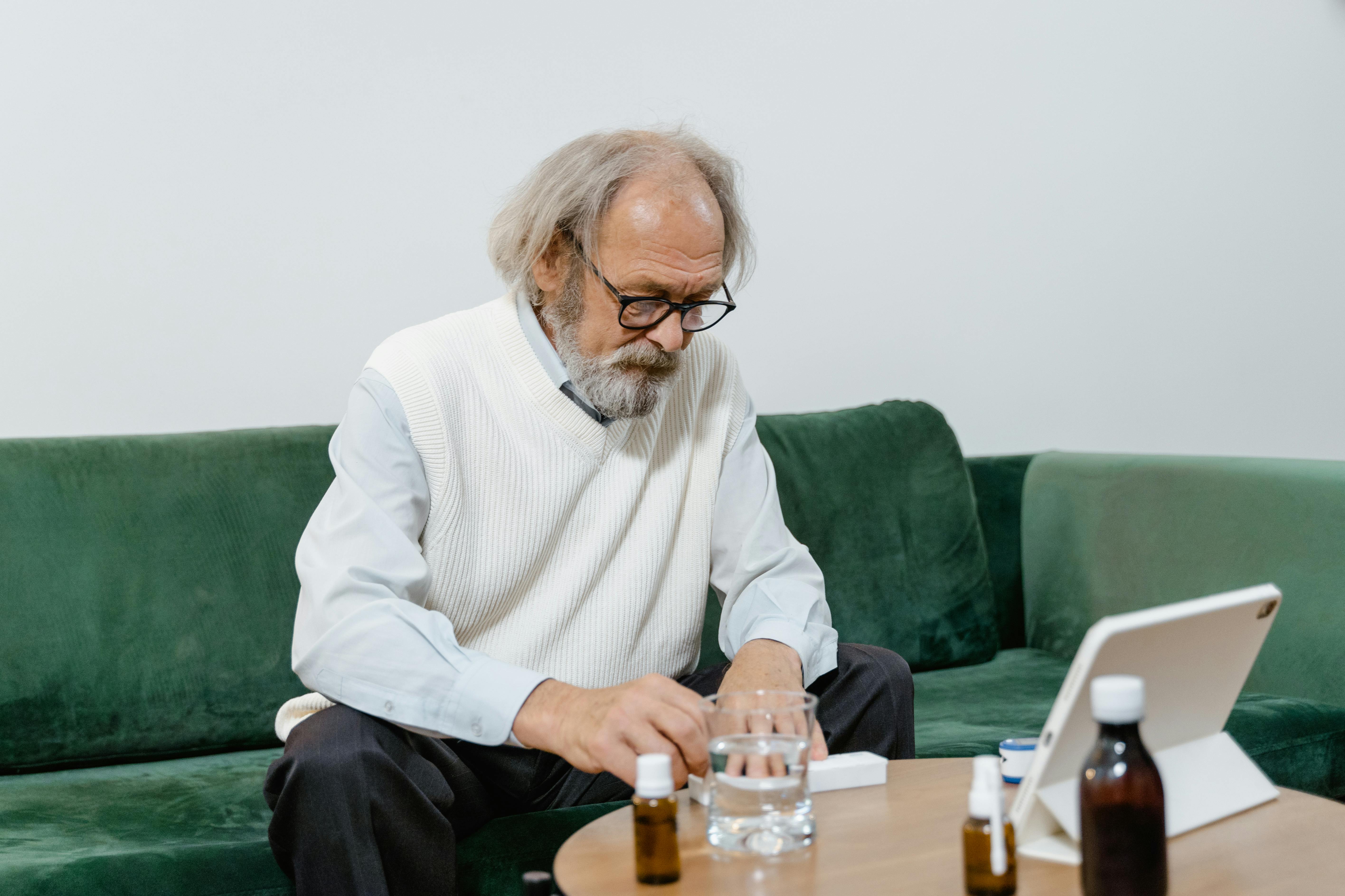 Ein älterer Mann sitzt vor einem Tablet und denkt nach. Auf dem Tisch stehen Medikamente und ein Glas Wasser.