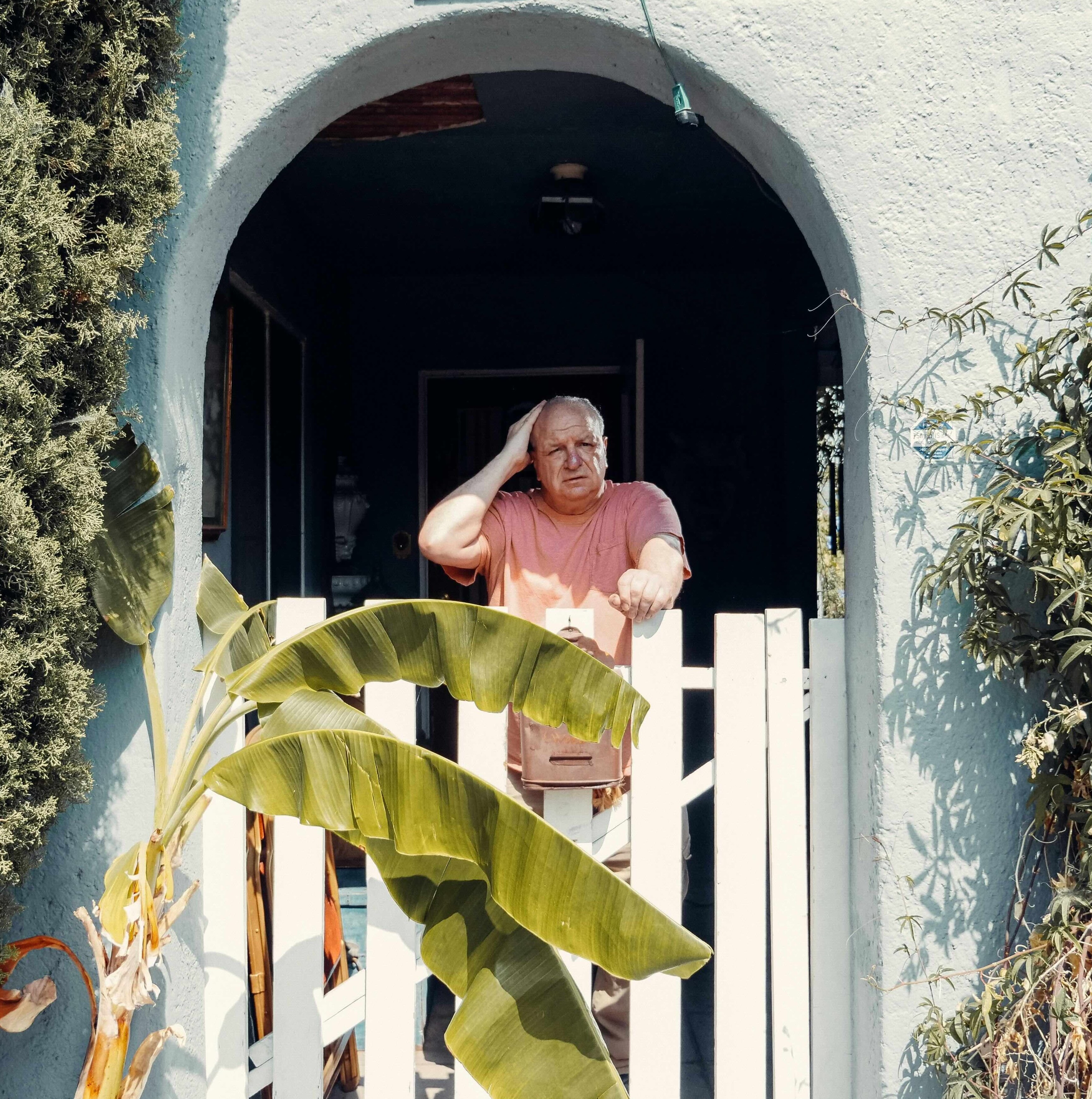 Ein älterer Mann steht nachdenklich vor seinem Haus und stützt sich auf ein Gartentor.