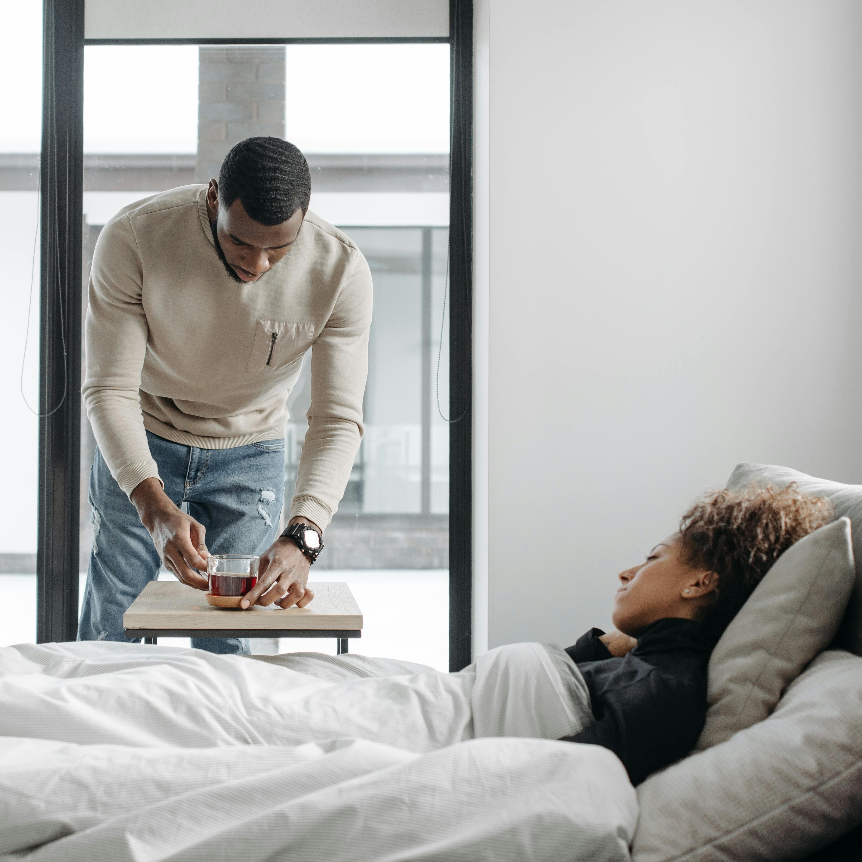 Ein Mann stellt einer im Bett liegenden Frau eine Tasse Tee ans Bett.