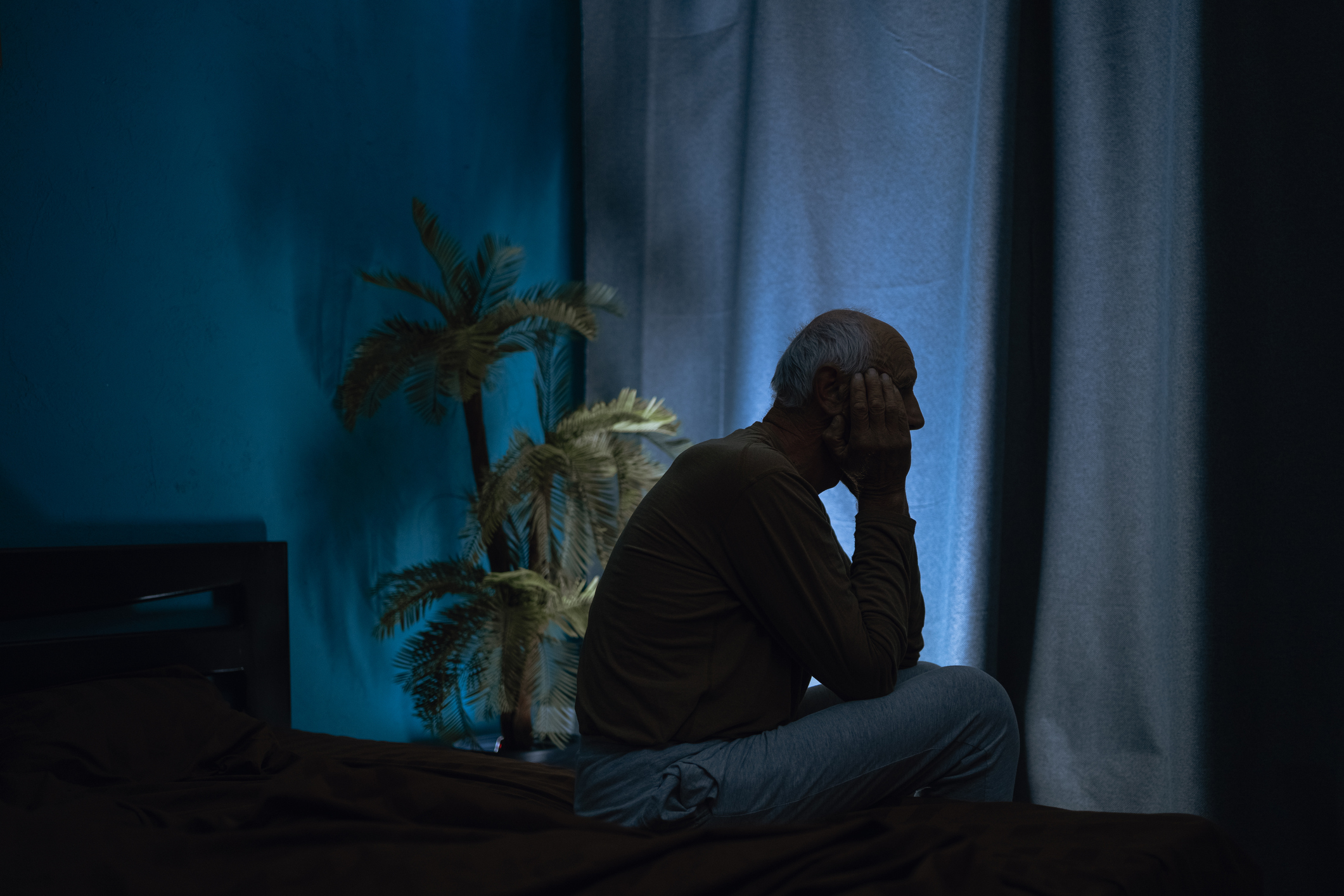 Ein älterer Mann sitzt im dunklen Schlafzimmer an der Bettkante. Er stütz den Kopf in den Arm.