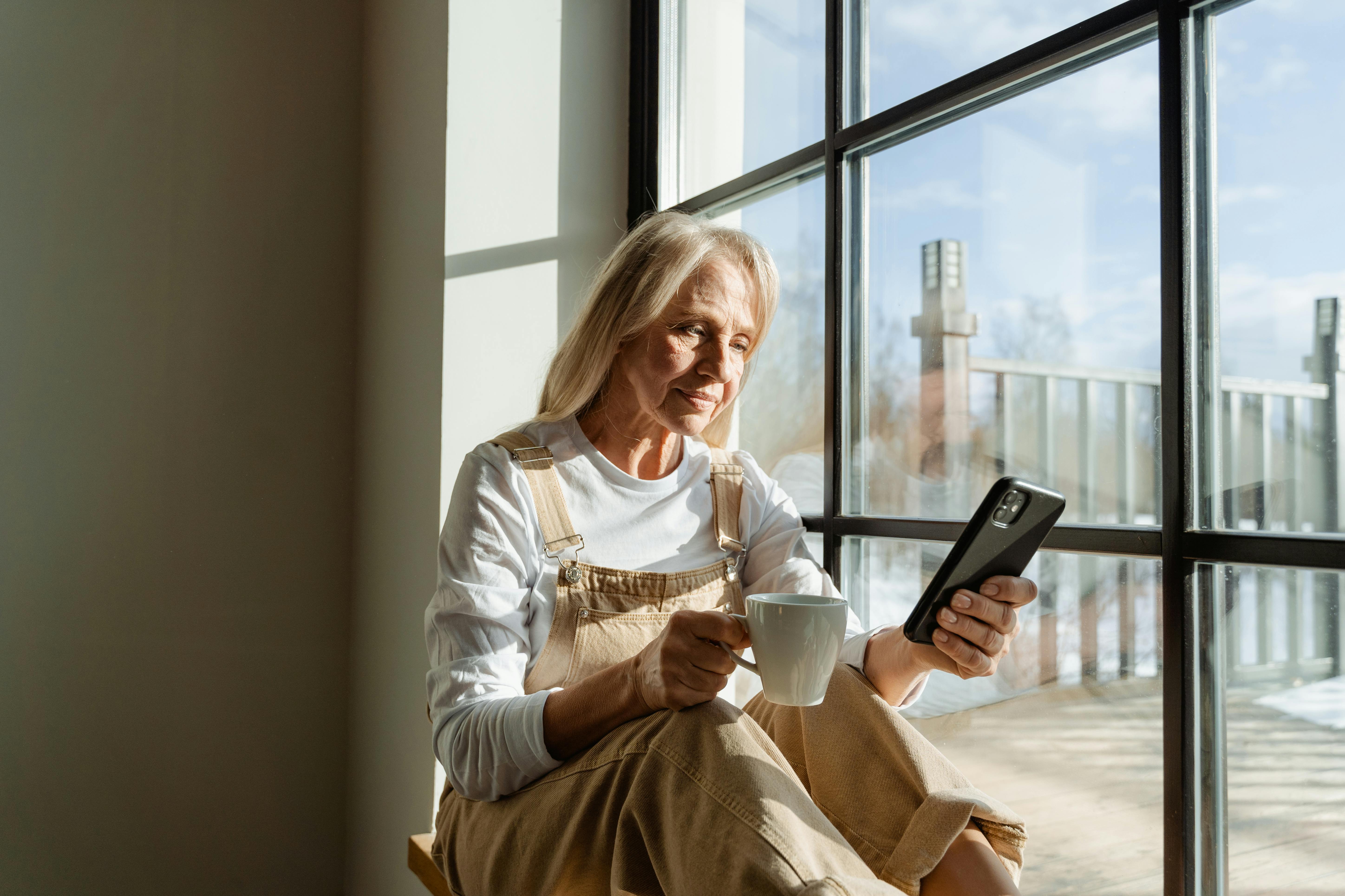 Eine Frau sitzt am Fenster. Sie trinkt Kaffee und schaut in ihr Mobiltelefon.