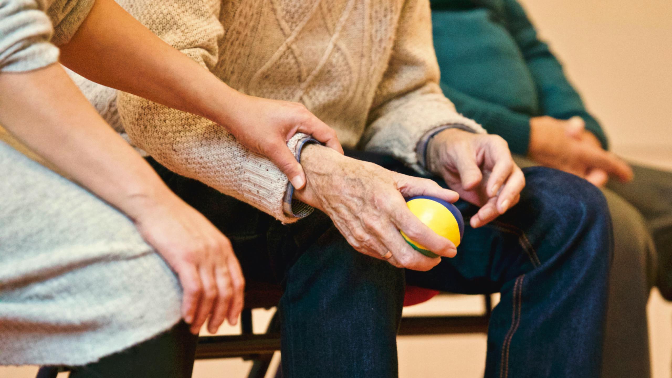 Pflegerin st&uuml;tzt die Hand eines &auml;lteren Mannes, der einen Igelball h&auml;lt