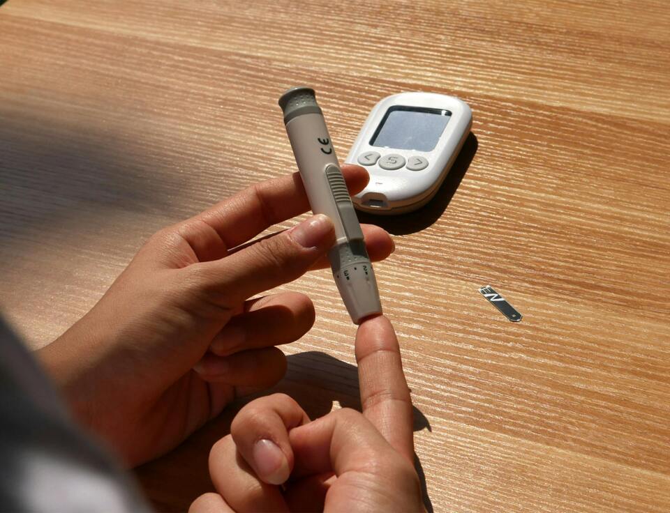 H&auml;nde setzen eine Stechhilfe zur Blutzuckermessung an, daneben ein Blutzuckermessger&auml;t
