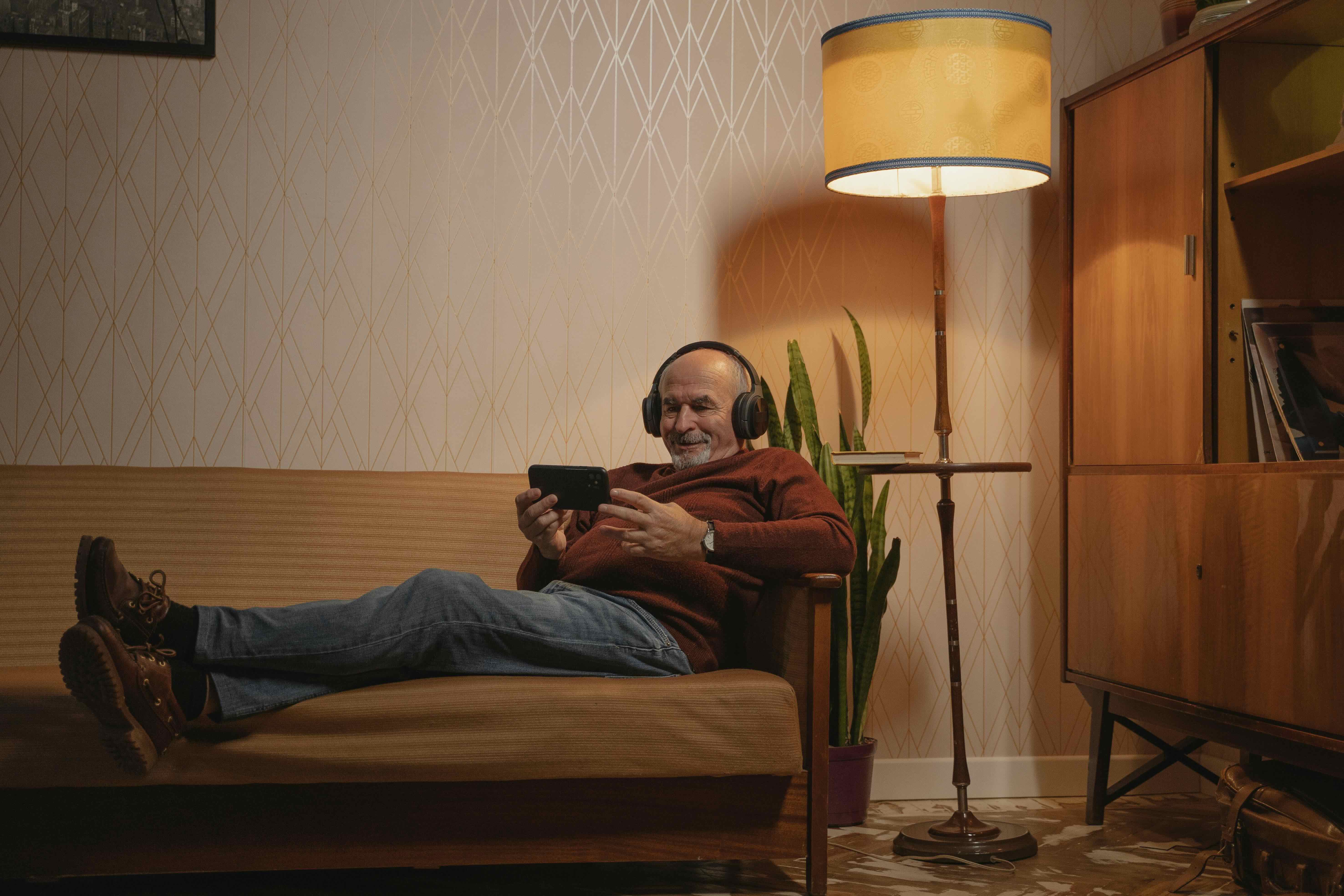 &Auml;lterer Mann mit Kopfh&ouml;rern entspannt auf dem Sofa und schaut auf sein Smartphone